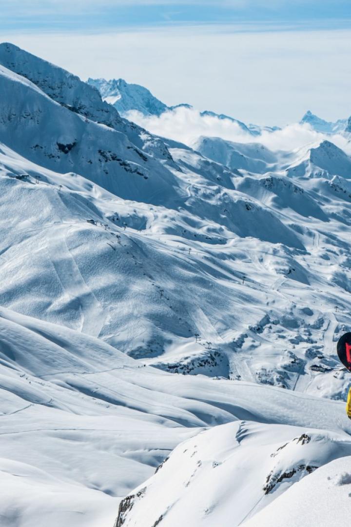 Zwei Snowboarder stehen auf einem verschneiten Gipfel im Skigebiet Arlberg und blicken auf die imposanten Alpen mit langen, leeren Abfahrten.