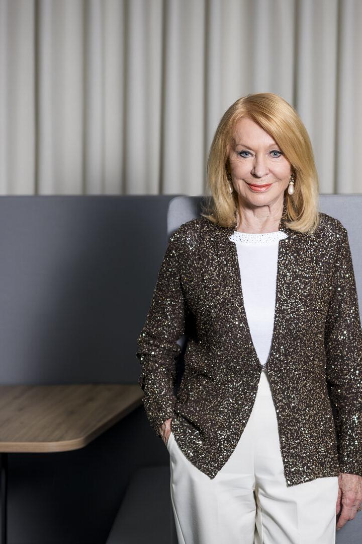 Eine Frau mit blondem Haar trägt eine glitzernde Jacke und weiße Hose, steht vor grauen Sitzmöbeln und hellem Vorhang.