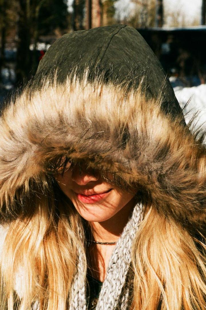 Eine Frau trägt eine Winterjacke mit Fellkapuze im Schnee, wobei das Fell außen sichtbar das Gesicht vor Kälte schützt.