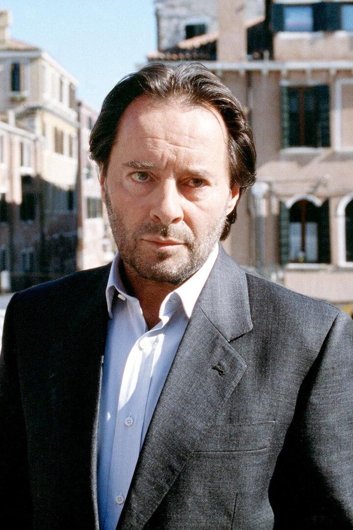 Ein Mann im Anzug steht auf einer Brücke vor alten Häusern mit venezianischer Architektur.