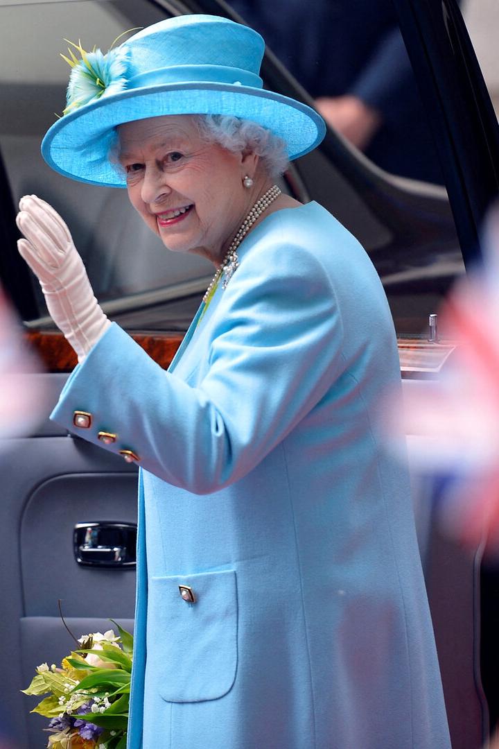Königin Elisabeth II. in hellblauem Kostüm und Hut winkt aus einem Auto.
