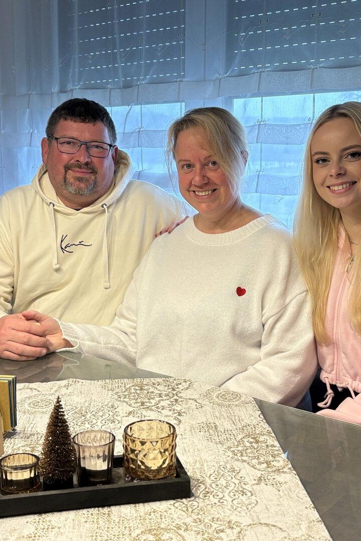 Drei Personen sitzen lächelnd an einem Tisch mit Kerzen und Dekoration, im Hintergrund ein Fenster mit Gardinen.