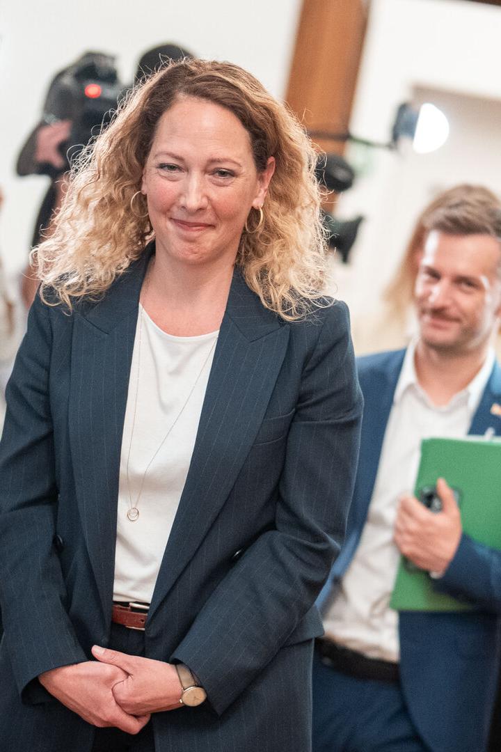 Eine Frau im Anzug geht lächelnd durch einen Flur, gefolgt von mehreren Personen mit Aktenmappen und umgeben von Fotografen.