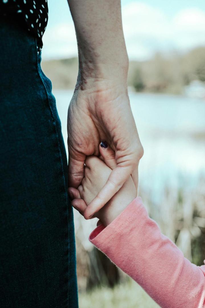 Ein Kind hält die Hand einer erwachsenen Person im Freien, beide stehen nebeneinander an einem Gewässer.