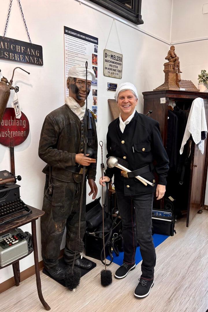 Eine Person in traditioneller Schornsteinfeger-Uniform steht lächelnd neben einer Schornsteinfeger-Figur mit Werkzeugen.