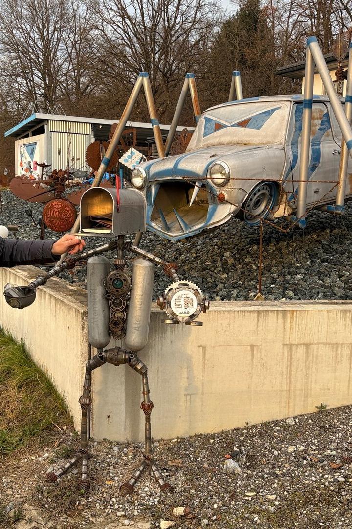 Ein Mann steht neben einer skurrilen Metallskulptur, im Hintergrund ein Auto mit Spinnenbeinen aus Metall.
