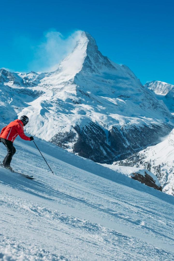 Eine Person fährt auf Skiern eine verschneite Piste hinab, im Hintergrund ragt das Matterhorn in den blauen Himmel.