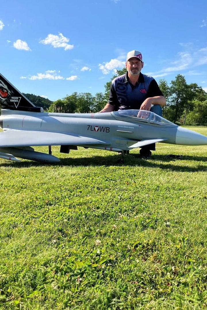 Ein Mann kniet auf einer Wiese hinter einem großen Modellflugzeug, umgeben von Bäumen und blauem Himmel.