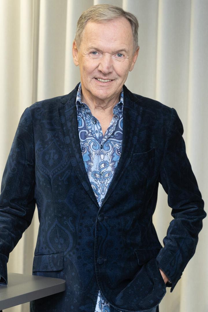 Ein lächelnder Mann mit blauem Hemd und Sakko steht vor einem hellen Vorhang.