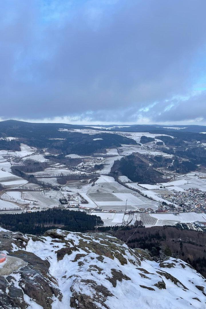 Verschneite Felder und Dörfer in einem weiten Tal, im Vordergrund Felsen mit etwas Schnee und kahlen Ästen.