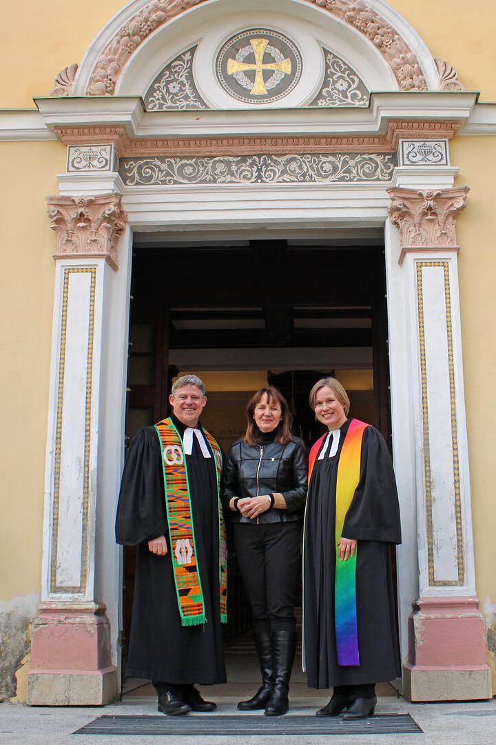 Drei Personen stehen vor dem Eingang einer Kirche, zwei davon tragen schwarze Talare mit bunten Stolen.