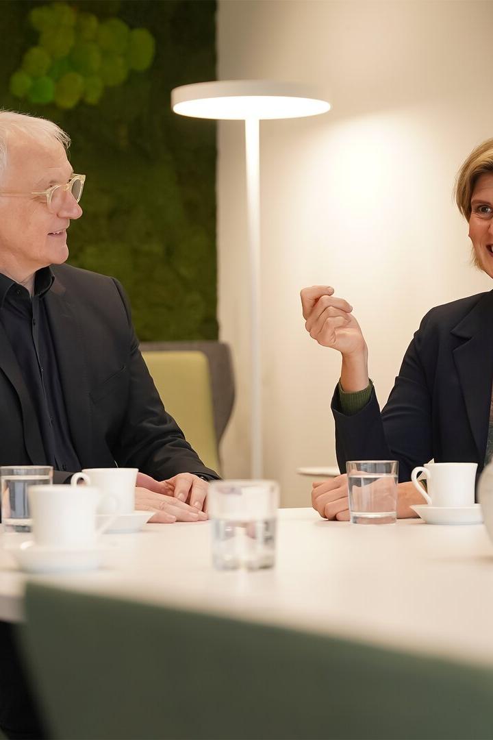 Zwei Personen im Gespräch an einem Tisch mit Tassen, Gläsern und silbernen Dekoräpfeln, beide wirken freundlich.