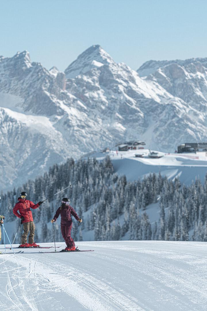Drei Skifahrer stehen auf einer verschneiten Piste vor einer Kulisse aus schneebedeckten Bergen.