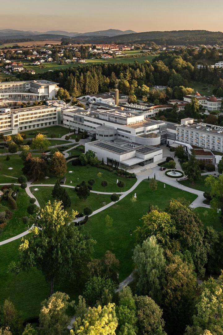 Mehrere moderne Gebäude mit weißen Fassaden liegen inmitten einer weitläufigen, grünen Parkanlage mit Wegen und Bäumen.