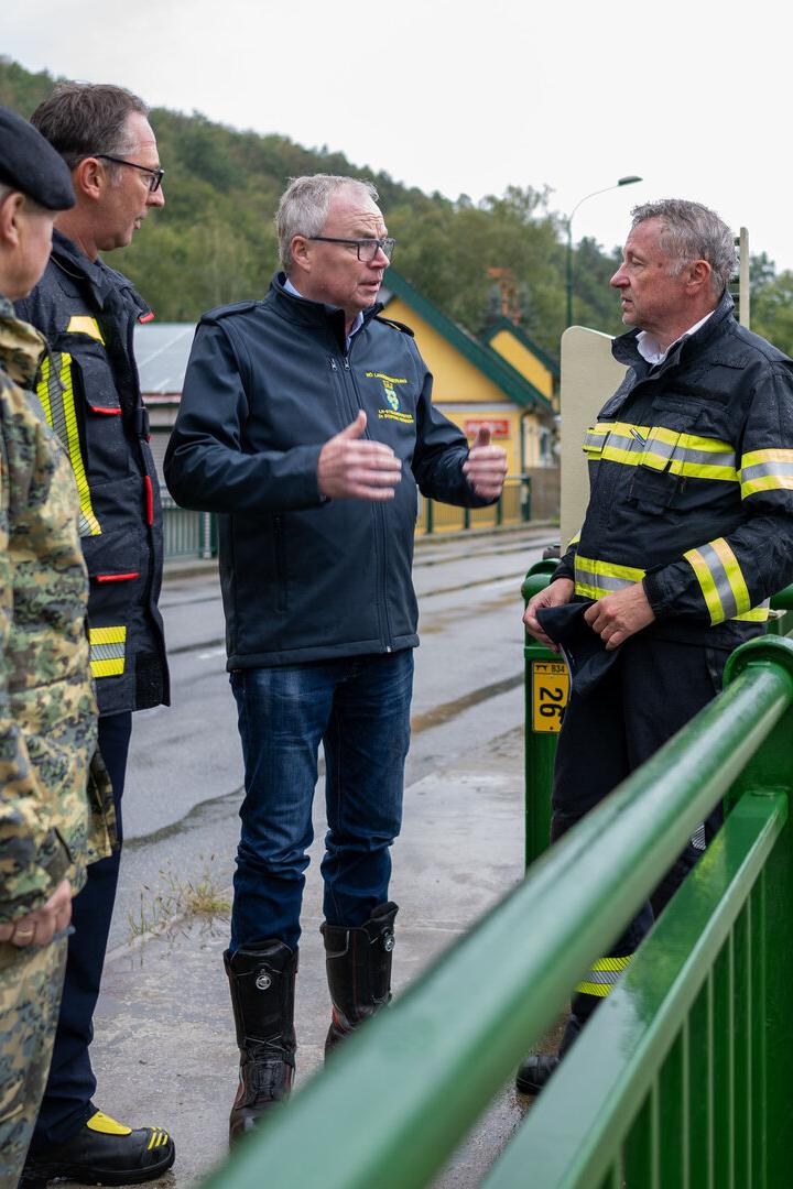 Vier Männer in Uniformen stehen auf einer Brücke und besprechen die Hochwassersituation am reißenden Fluss.