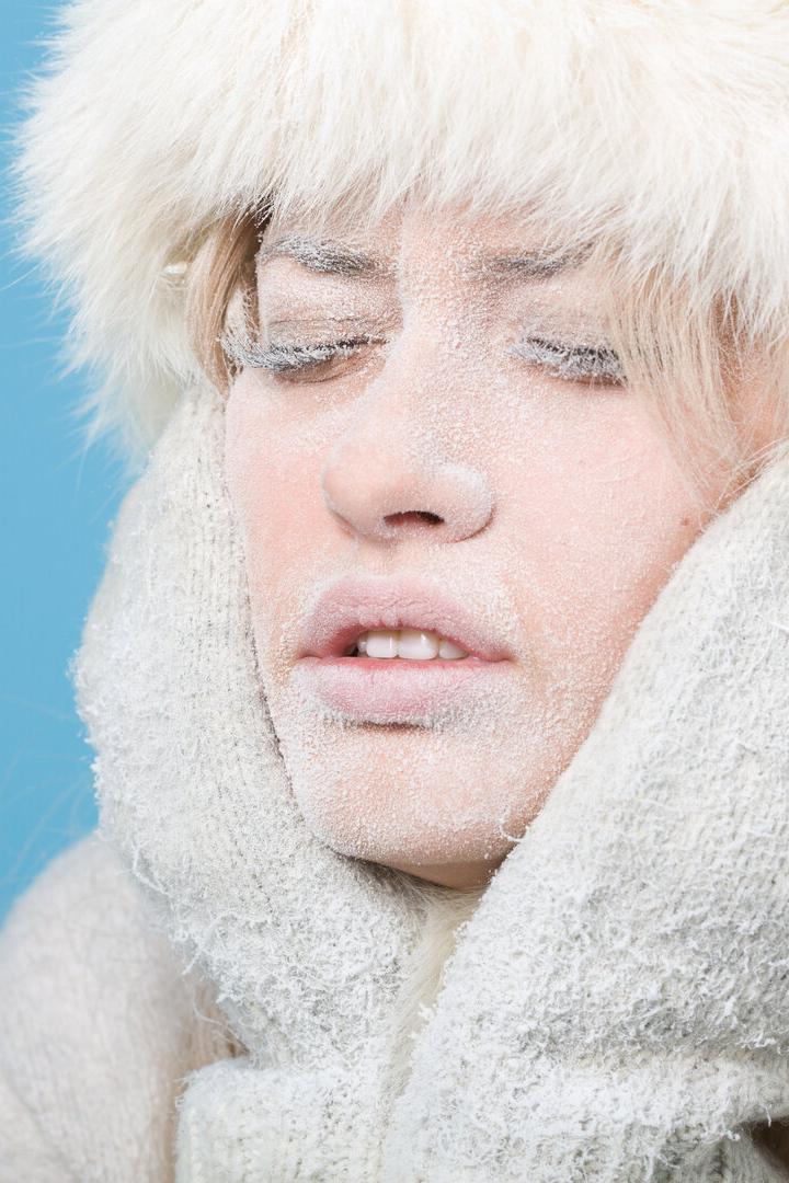 Eine Frau mit frostbedecktem Gesicht trägt eine Pelzmütze und Handschuhe vor blauem Hintergrund.