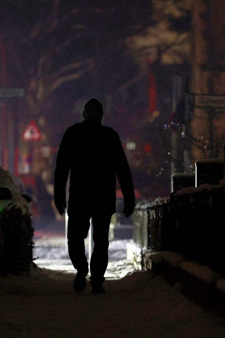 Eine Person geht nachts auf einem verschneiten Gehweg zwischen parkenden Autos und Häusern.