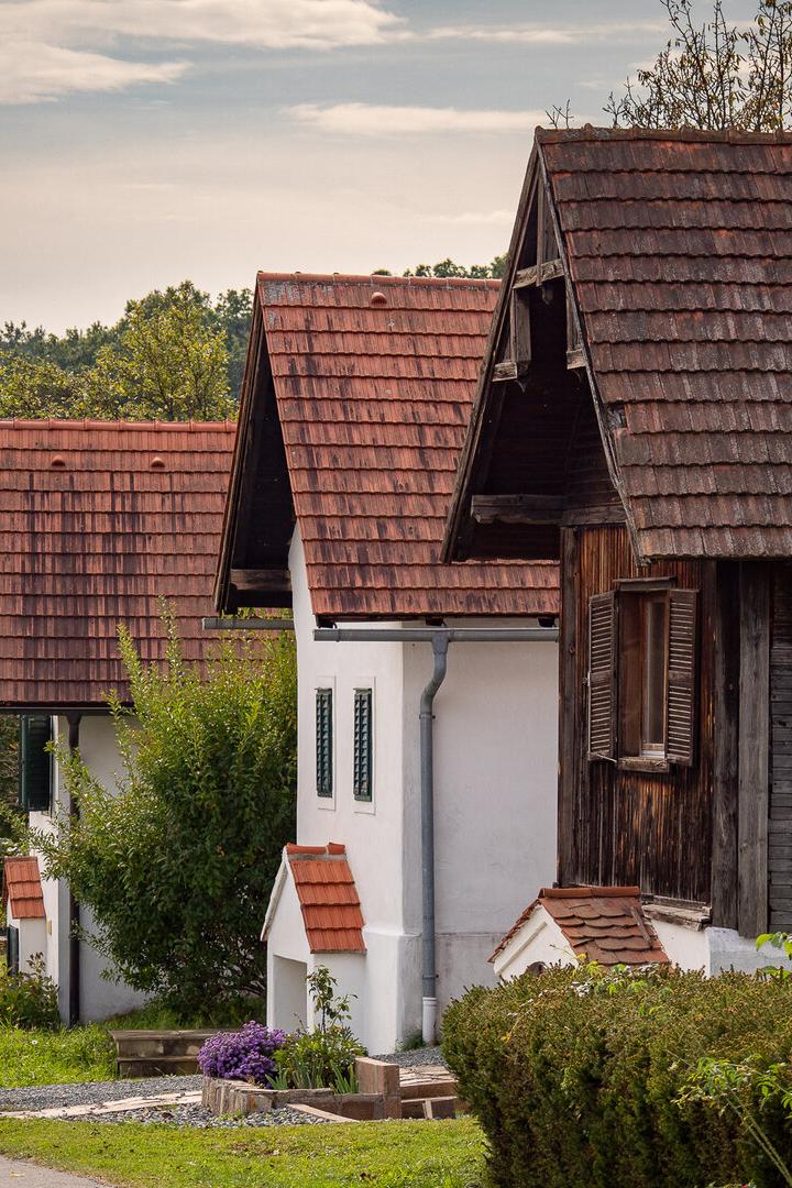 Mehrere kleine Häuser mit roten Ziegeldächern stehen nebeneinander an einer ruhigen Straße, umgeben von viel Grün.