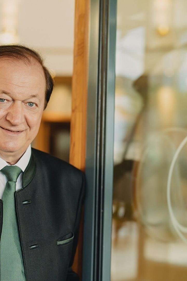 Ein älterer Mann im Trachtenanzug mit grünem Schlips steht lächelnd neben einer Glastür mit verziertem Logo.