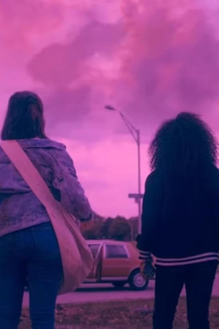 Vier Jugendliche stehen nebeneinander und blicken unter einem pink-violetten Himmel auf eine Straße mit geparktem Auto.