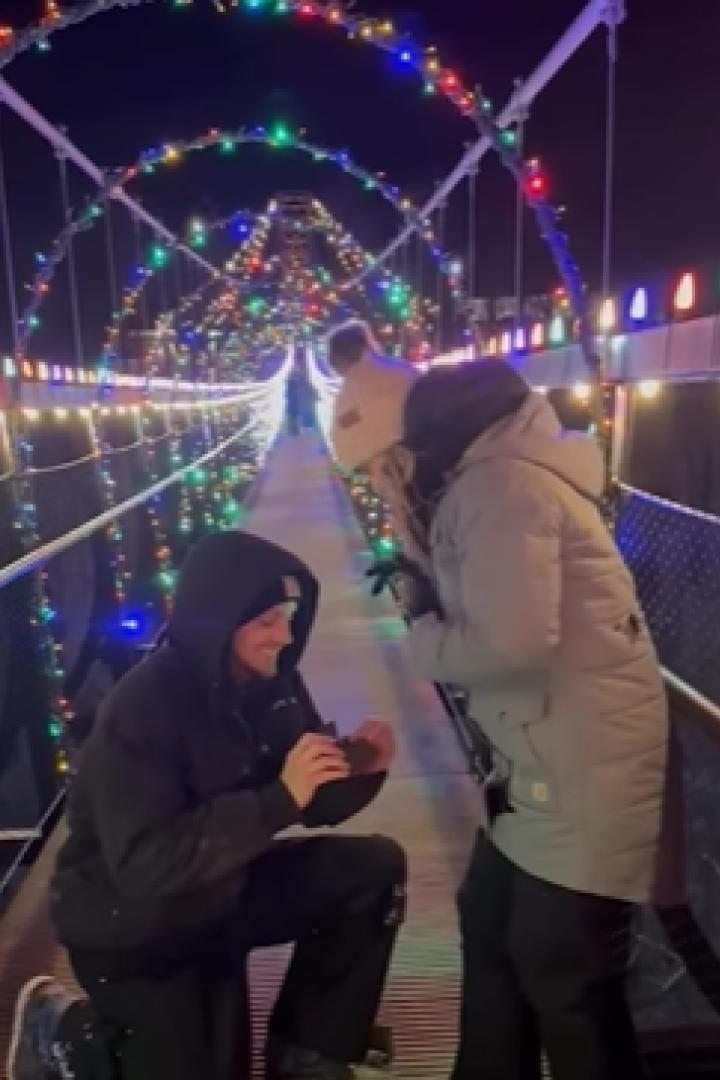 Mann kniet auf beleuchteter Hängebrücke und hält einer Frau einen Ring entgegen.