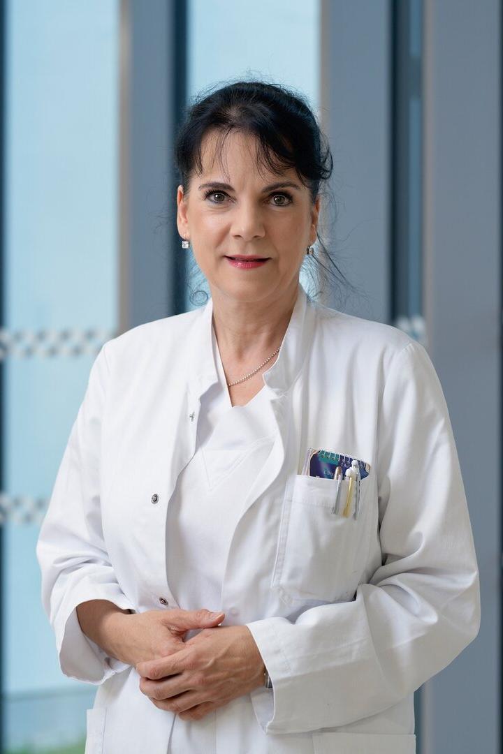 Eine Frau im weißen Arztkittel steht mit Stiften in der Brusttasche vor großen Fenstern und blickt freundlich nach vorn.