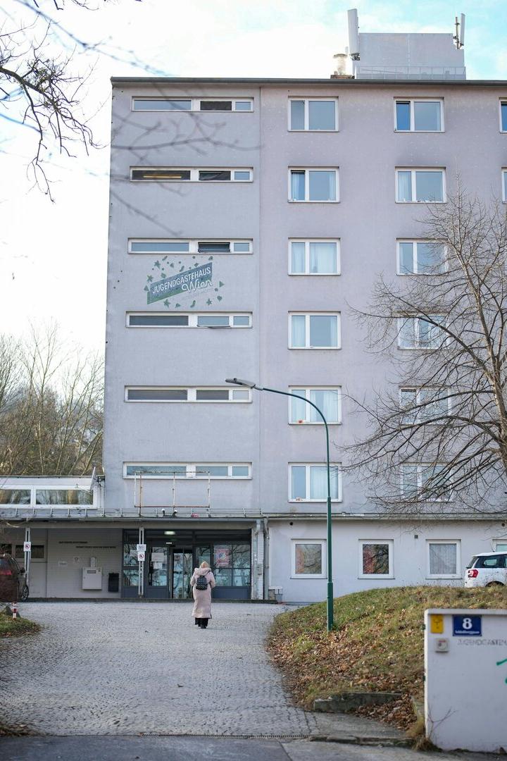 Ein mehrstöckiges graues Gebäude mit vielen Fenstern, davor eine Person auf einem gepflasterten Weg und kahle Bäume.