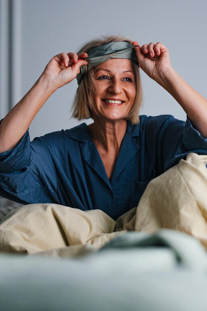 Eine lächelnde Frau im blauen Pyjama sitzt im Bett und zieht sich eine Schlafmaske vom Kopf.