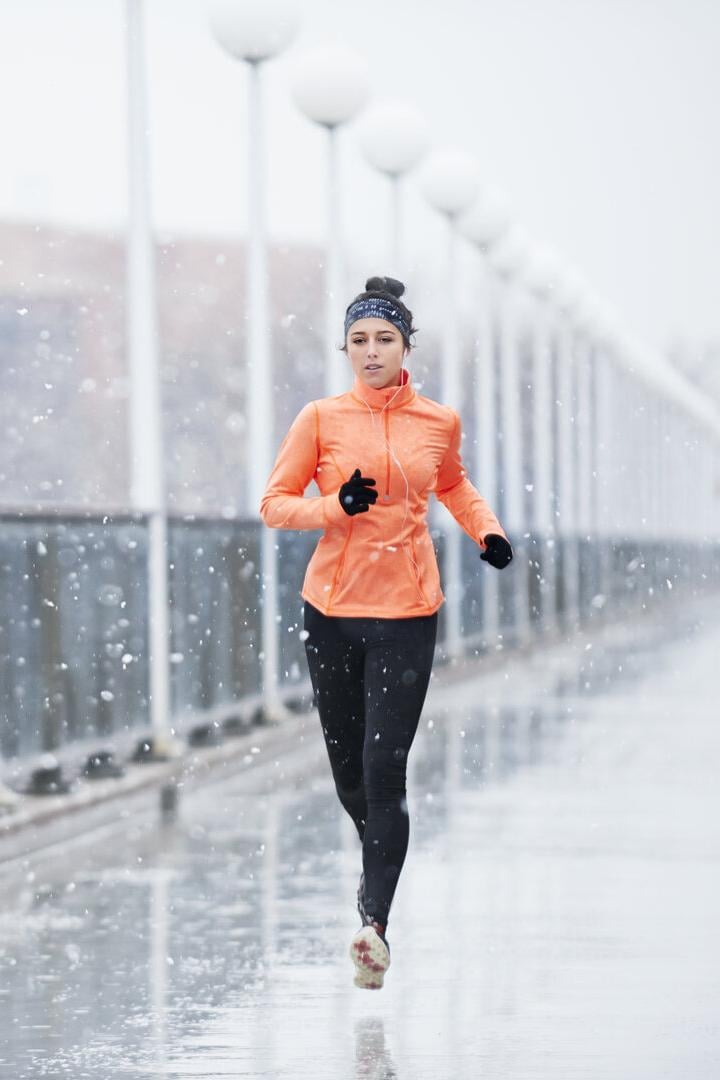 Eine Frau joggt bei Schneefall auf einer Brücke und trägt eine orange Jacke sowie schwarze Leggings.