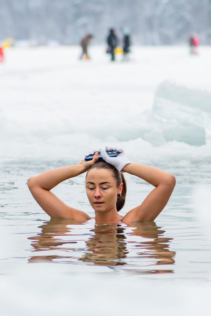 Eine Frau badet in einem eisigen See, umgeben von Schnee, und hält ihre Hände mit Handschuhen auf dem Kopf.