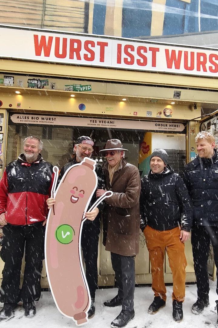 Mehrere Menschen stehen vor dem Würstelstand mit Aufschrift "Wurst isst Wurst"