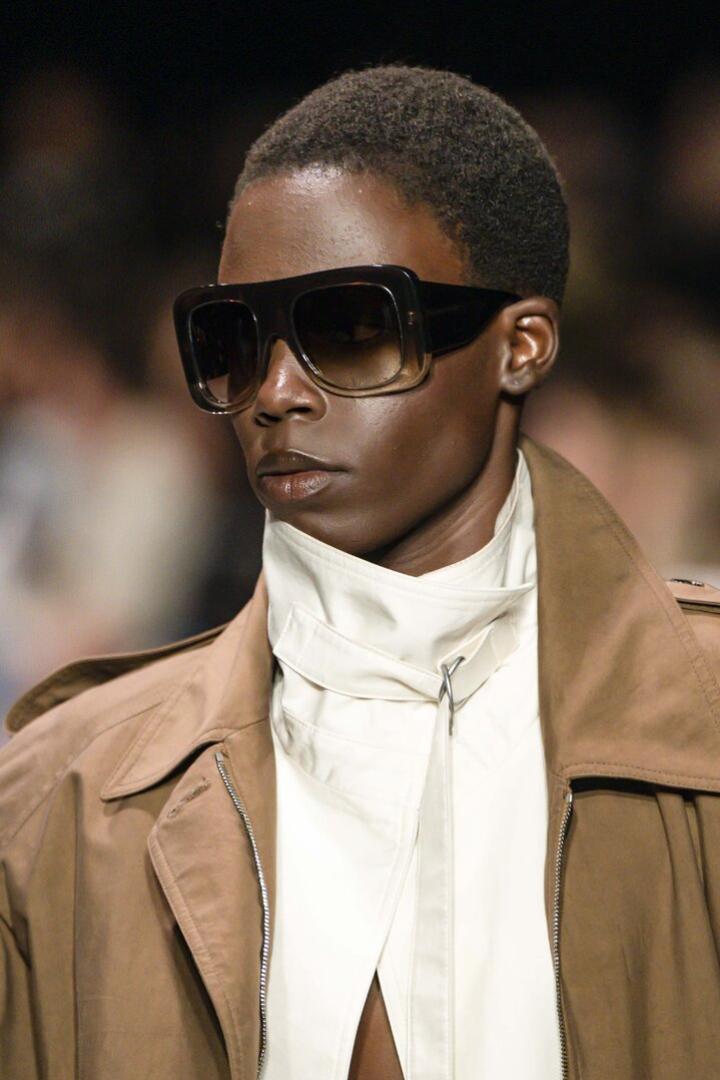 Person mit großer, dunkler Sonnenbrille, kurzem Haar, beigem Mantel und weißem, hochgeschlossenem Oberteil bei der Pariser Fashion Week