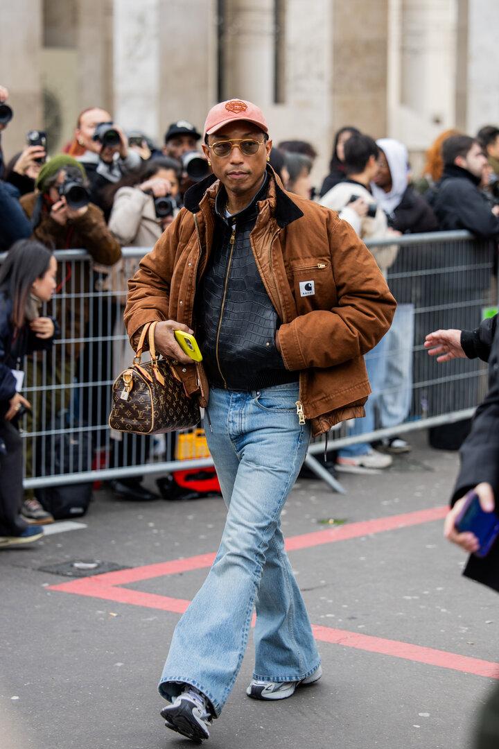 Pharrell Williams trägt eine braune Jacke, Schlaghosen und eine Louis Vuitton Tasche.