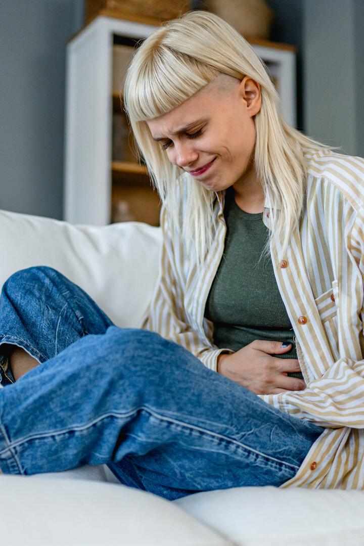 Eine junge Frau sitzt auf einem Sofa und hält sich mit schmerzverzerrtem Gesicht den Bauch.