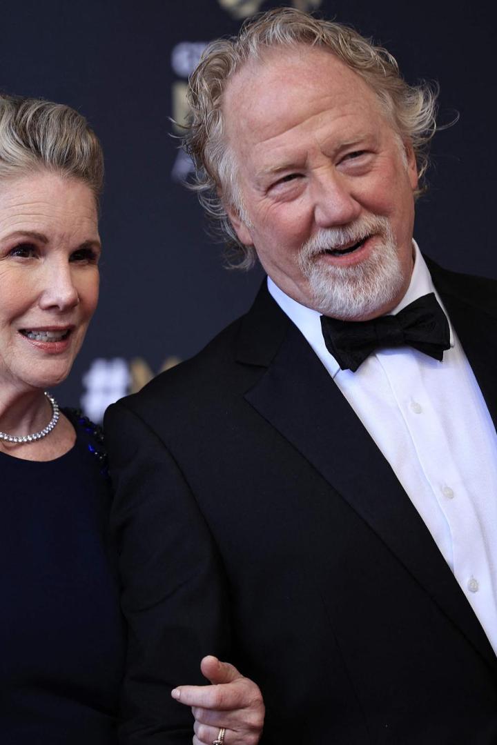 Ein Mann im Smoking mit Fliege und eine Frau im Abendkleid posieren lächelnd vor dunklem Hintergrund.