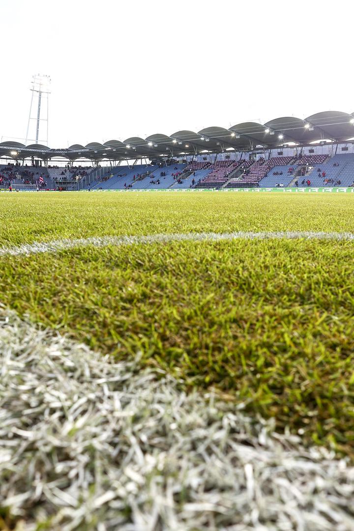 Ein Fußballfeld mit Rasen und weißen Linien vor einer Tribüne mit Zuschauern.
