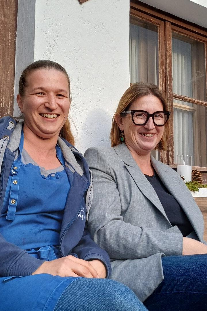 Die Almwirtinnen Agi und Michi sitzen auf einer Bank vor ihrem Gasthaus. 