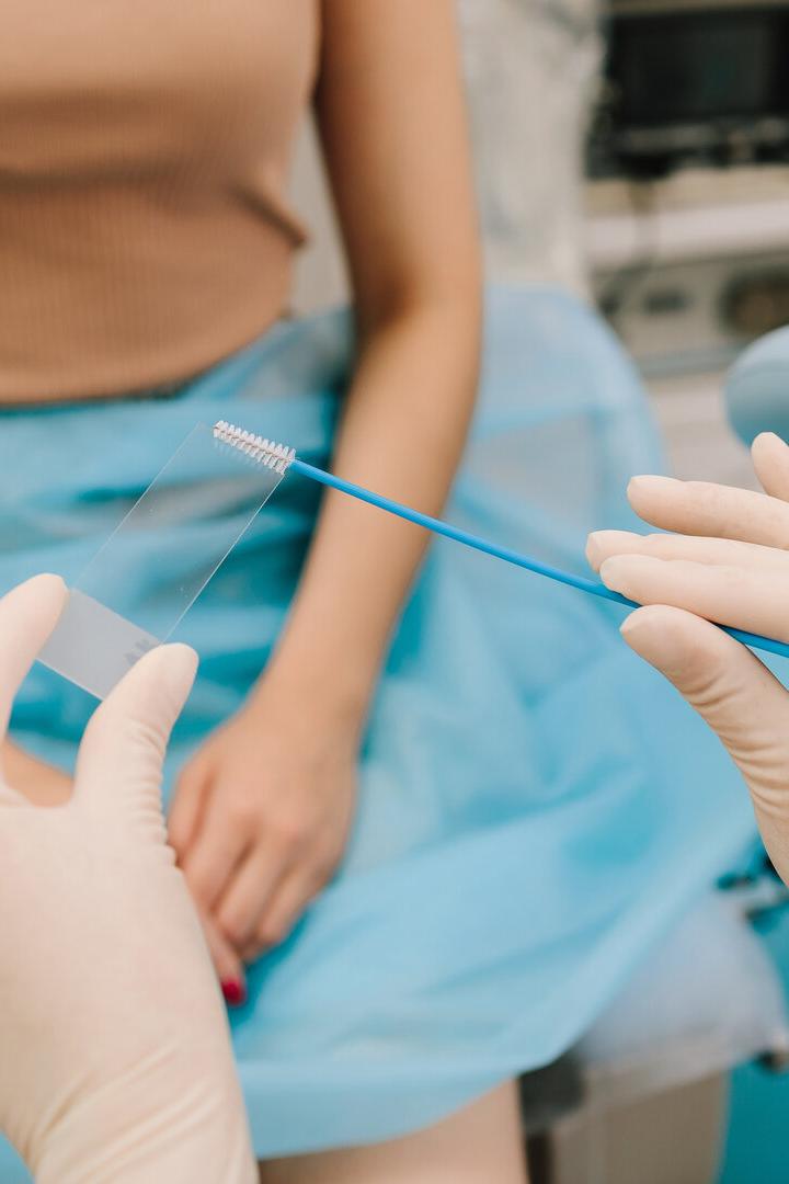 Eine Person mit Handschuhen hält einen Abstrich und einen Objektträger vor einer sitzenden Frau in medizinischer Umgebung.