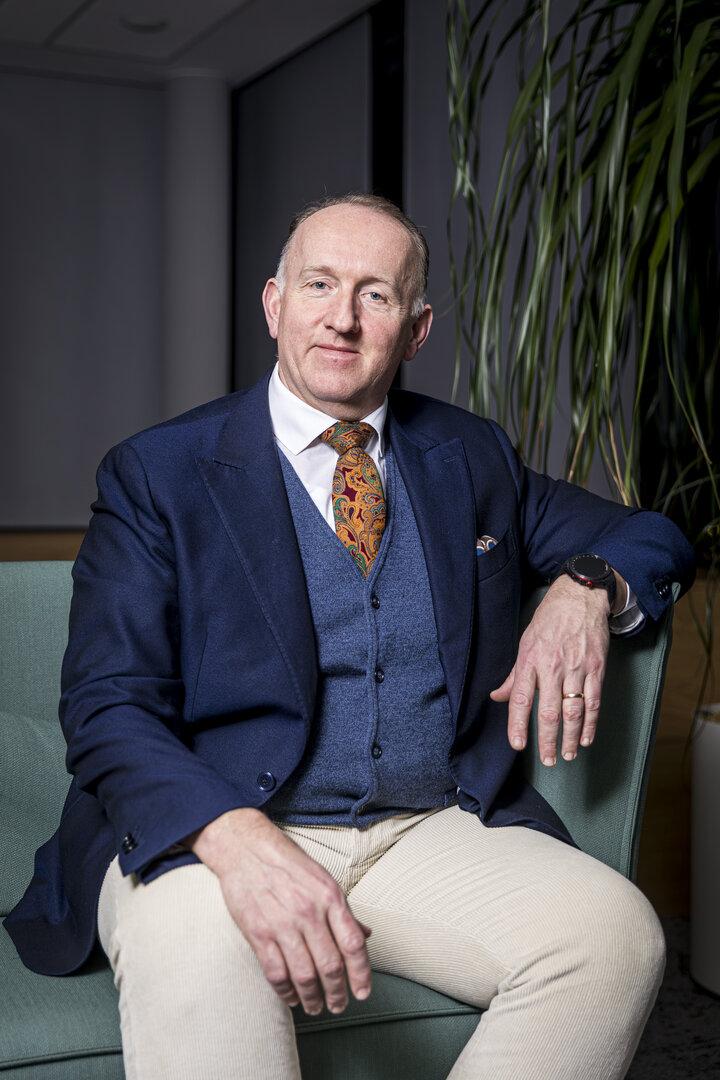 Ein Mann im Anzug mit bunter Krawatte sitzt auf einem grünen Sessel, im Hintergrund steht eine große Pflanze.