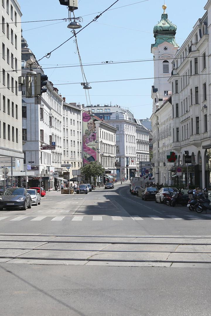 Mehrspurige Straße mit Autos, Straßenbahnschienen, Geschäften, Menschen auf Gehwegen und einer Kirche mit grünem Turm im Hintergrund.