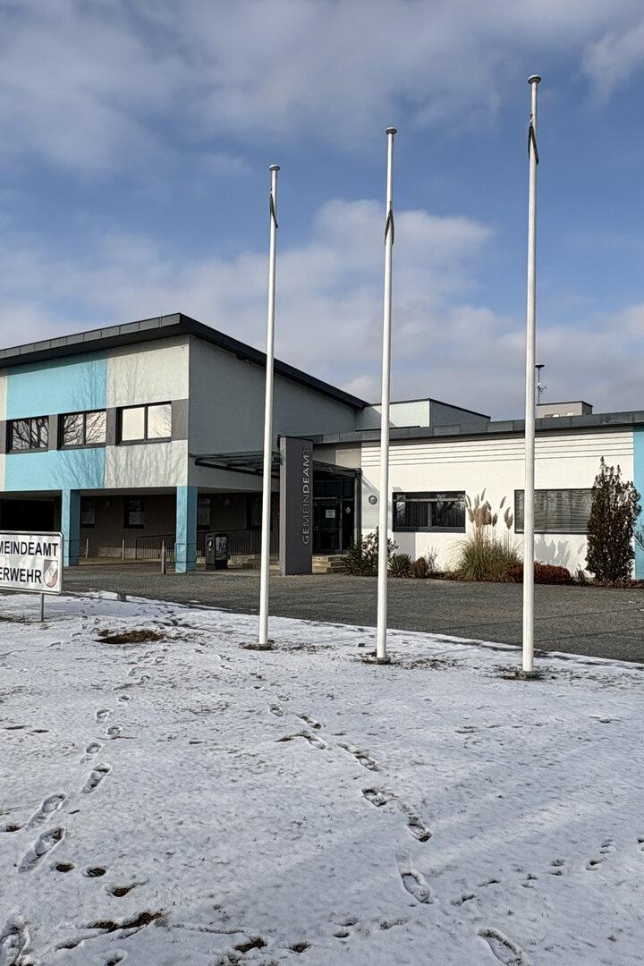 Ein Gebäude des Gemeindeamts/Feuerwehr mit vier Fahnenmasten steht vor einem teilweise schneebedeckten Feld.