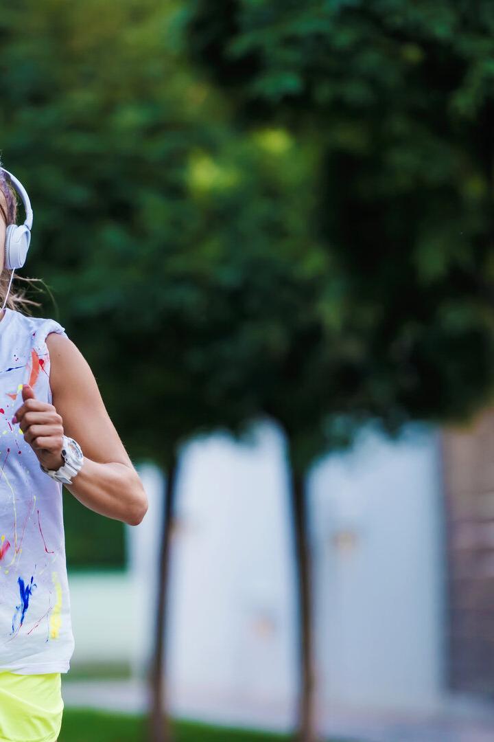 Junge Frau joggt mit Kopfhörern und bunter Sportkleidung auf einer Straße mit Bäumen im Hintergrund.