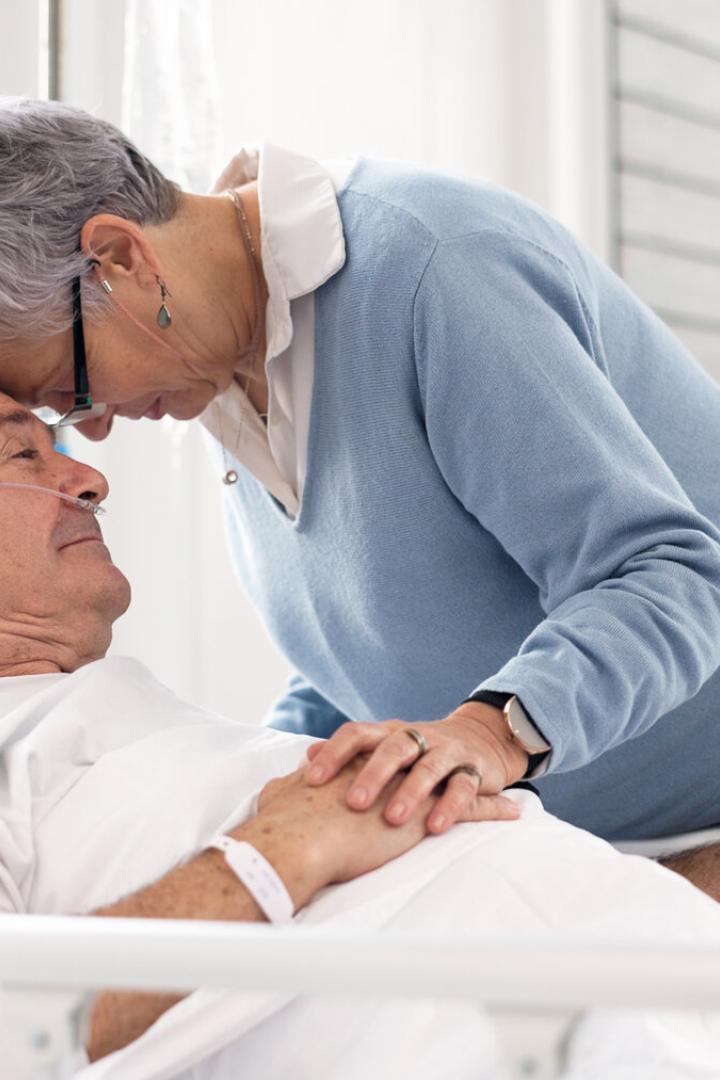 Eine ältere Frau hält liebevoll die Hand eines Mannes im Krankenhausbett und legt ihre Stirn an seine.