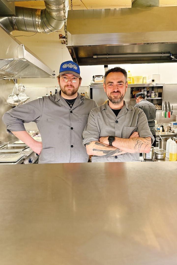 Zwei Köche in grauen Jacken stehen nebeneinander in einer professionellen Restaurantküche.