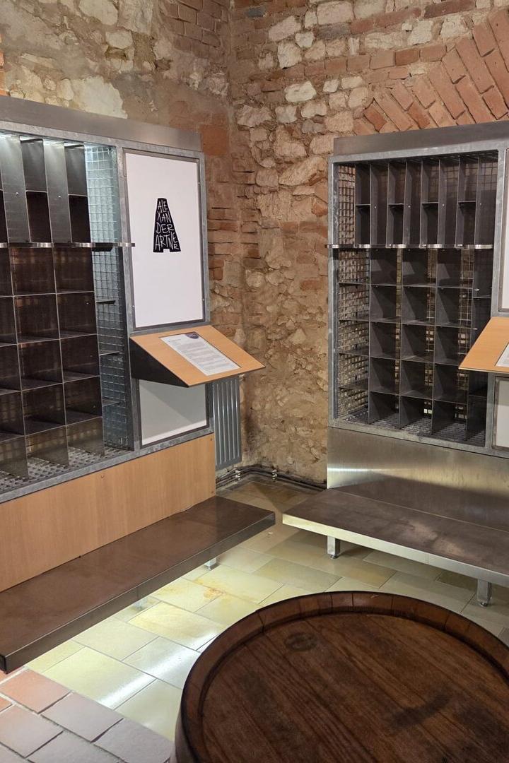 Zwei Metallregale mit nummerierten Fächern und Infotafeln stehen vor einer Backsteinwand in einem historischen Raum.