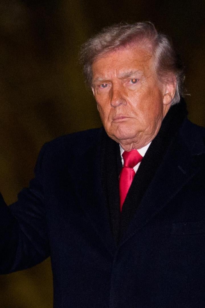 Donald Trump in dunklem Mantel und roter Krawatte grüßt mit einem schwarzen Handschuh.