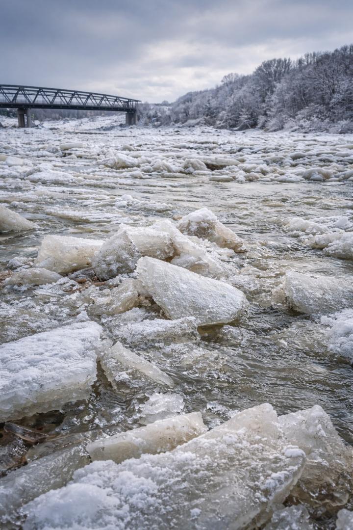 Eine Aufnahmen der von kleinen Eisschollen bedeckten Oberfläche eines Flusses. Im Hintergrund ist eine Brücke erkennbar.
