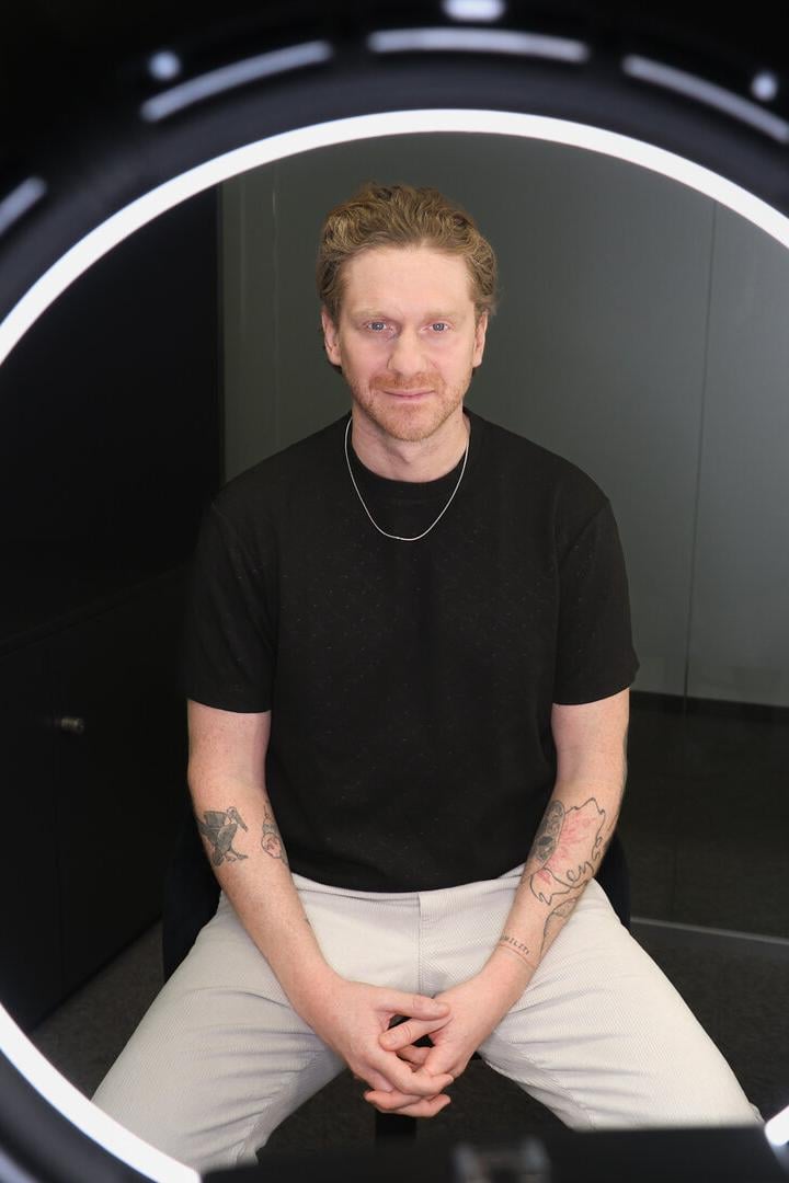 Ein Mann mit Tattoos sitzt vor einer leuchtenden Ringlampe und schaut direkt nach vorne.