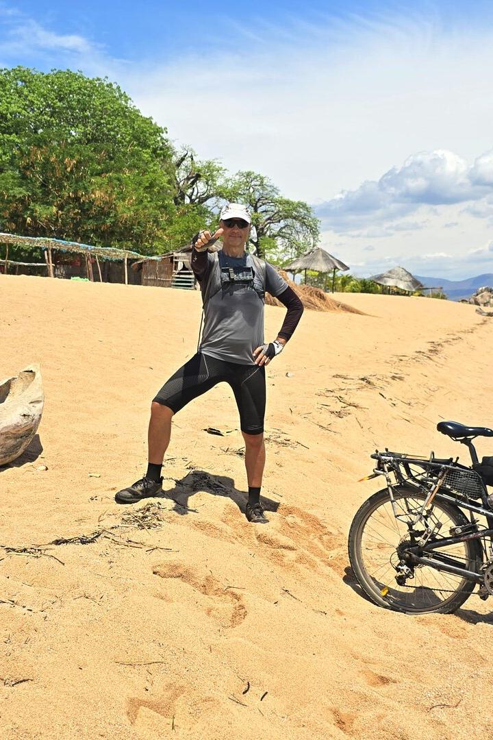 Eine Person steht auf einem Sandstrand neben einem Fahrrad und einem Einbaum, im Hintergrund Bäume und Wasser.