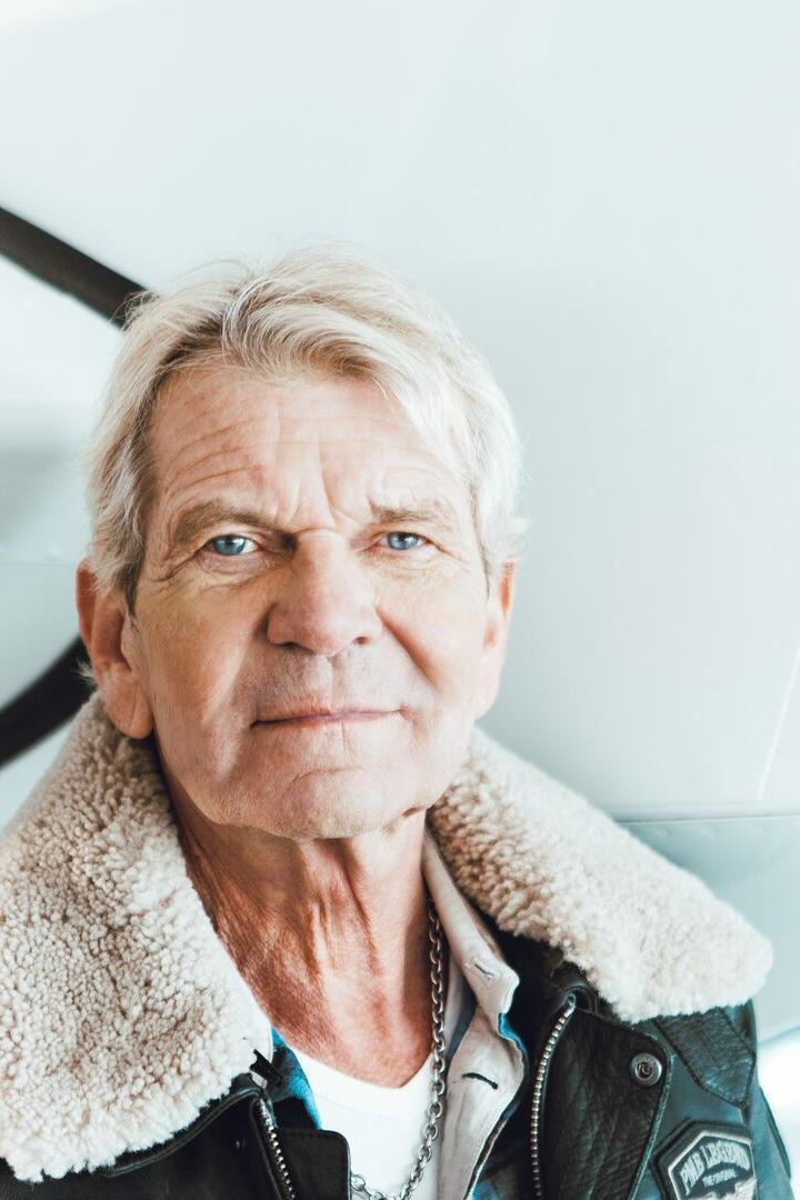Ein älterer Mann mit grauen Haaren und Fliegerjacke steht vor einem weißen Flugzeug.